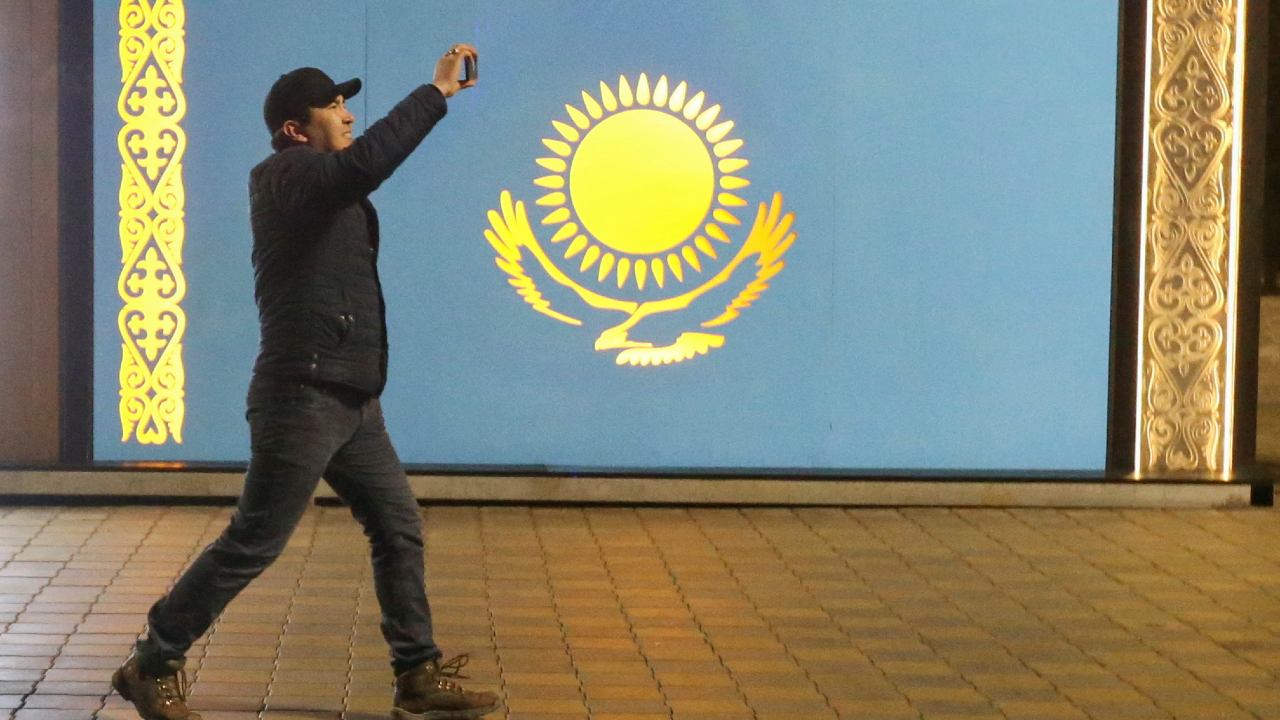 Photo: A man holds a mobile phone while walking past a board with a Kazakh state flag during a protest against LPG cost rise following authorities' decision to lift price caps on liquefied petroleum gas in Almaty, Kazakhstan January 5, 2022. Credit: REUTERS/Pavel Mikheyev